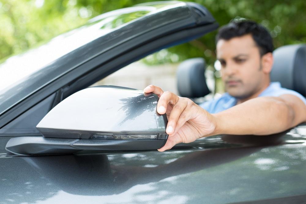 Closeup portrait, young man driver looking adjusting side view car mirror, making sure he can see traffic OK around him, isolated outdoors background