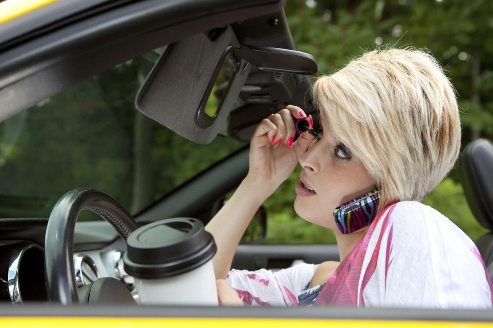 woman driving car while applying mascara, talking on her phone, and holding a coffee