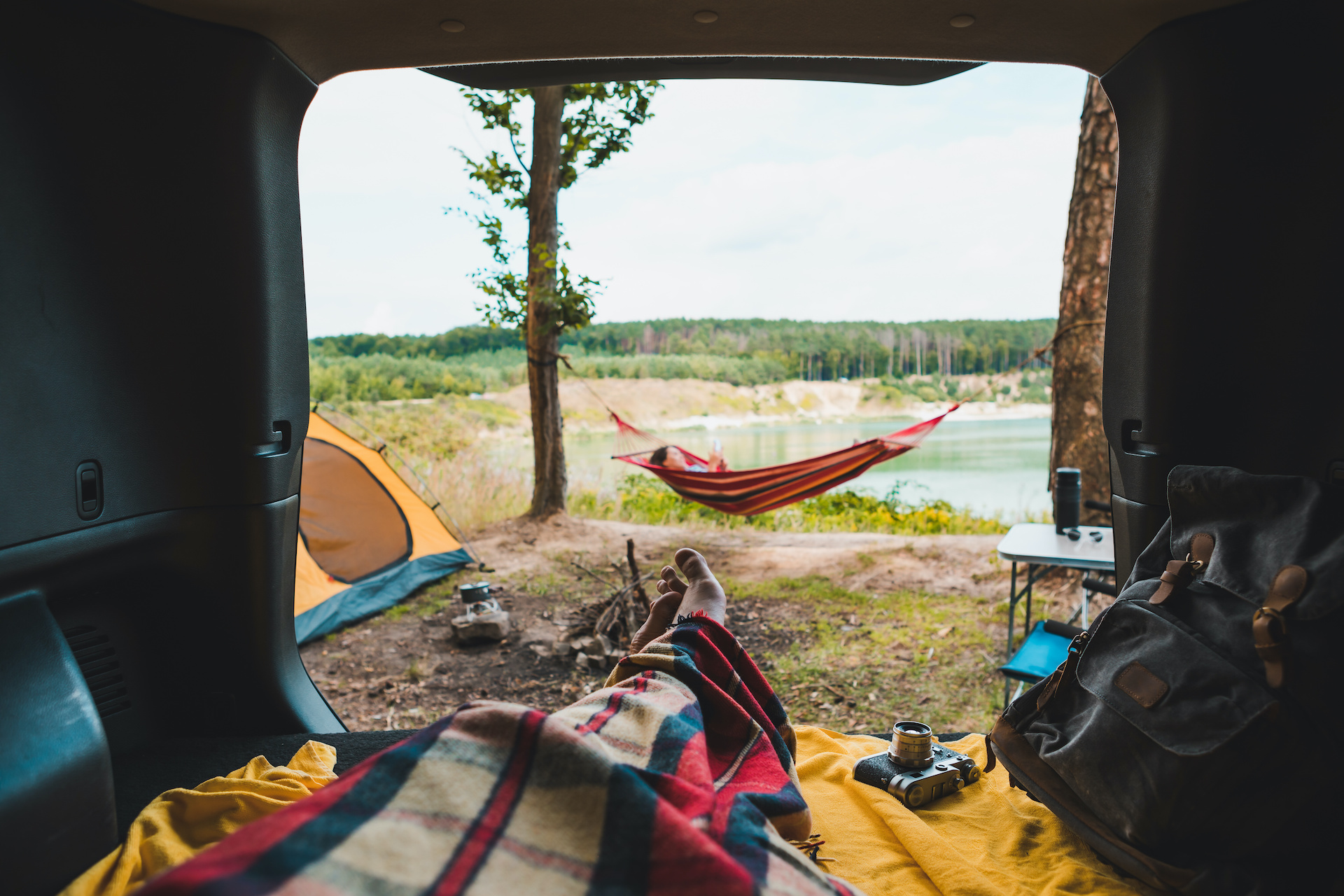 Person camping out of their car, watching another camper lay in a hammock and read.